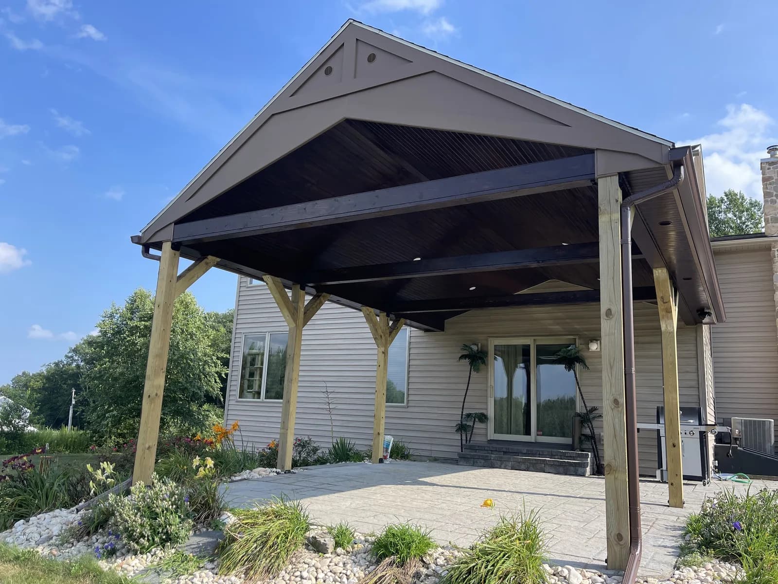 Gable-roof pavilion over a patio with planted garden edges beside a house.