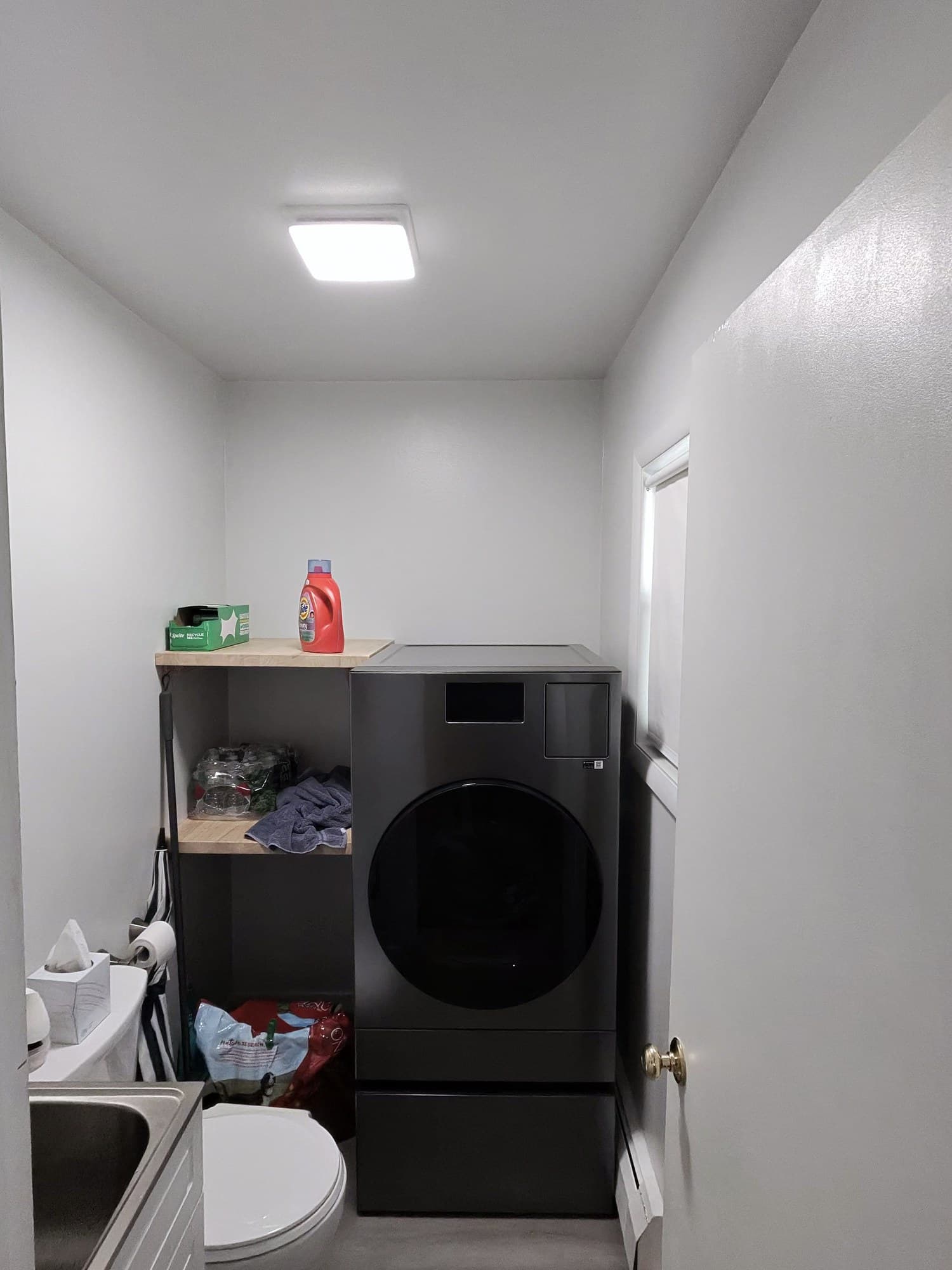 Full view of the remodeled laundry bathroom with washer, open shelves, and LED ceiling light.
